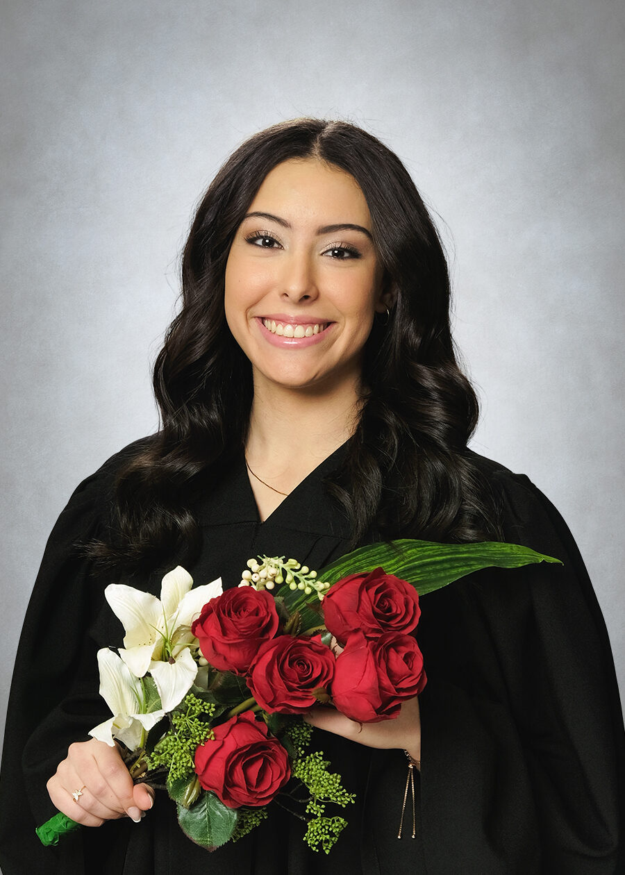 A high school graduation portrait of a girl wearing a graduation gown and holding flowers against a traditional grey background.