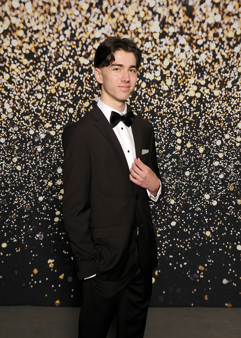 A high school prom photo of a boy wearing a tuxedo against a black and gold bokeh backgdrop.