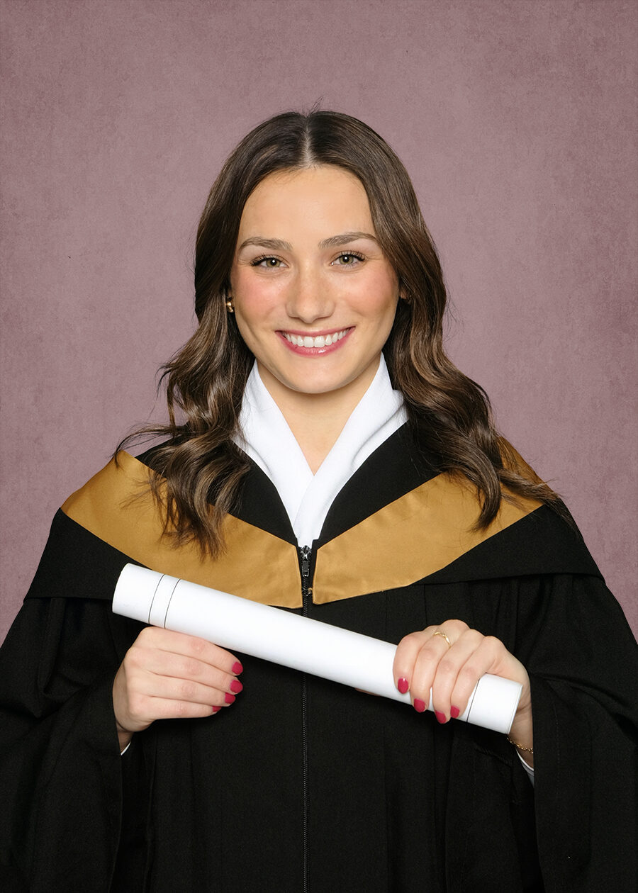University portrait of a young woman smiling wearnig a graduation gown holding a diploma against a pink traditional masters background