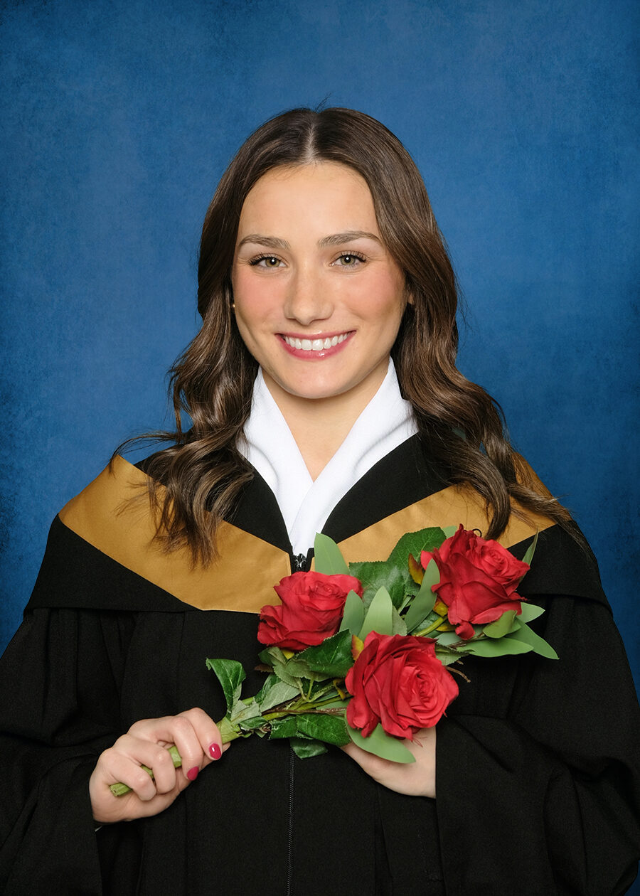 University portrait of a young woman smiling wearnig a graduation gown holding flowers against a blue traditional masters background
